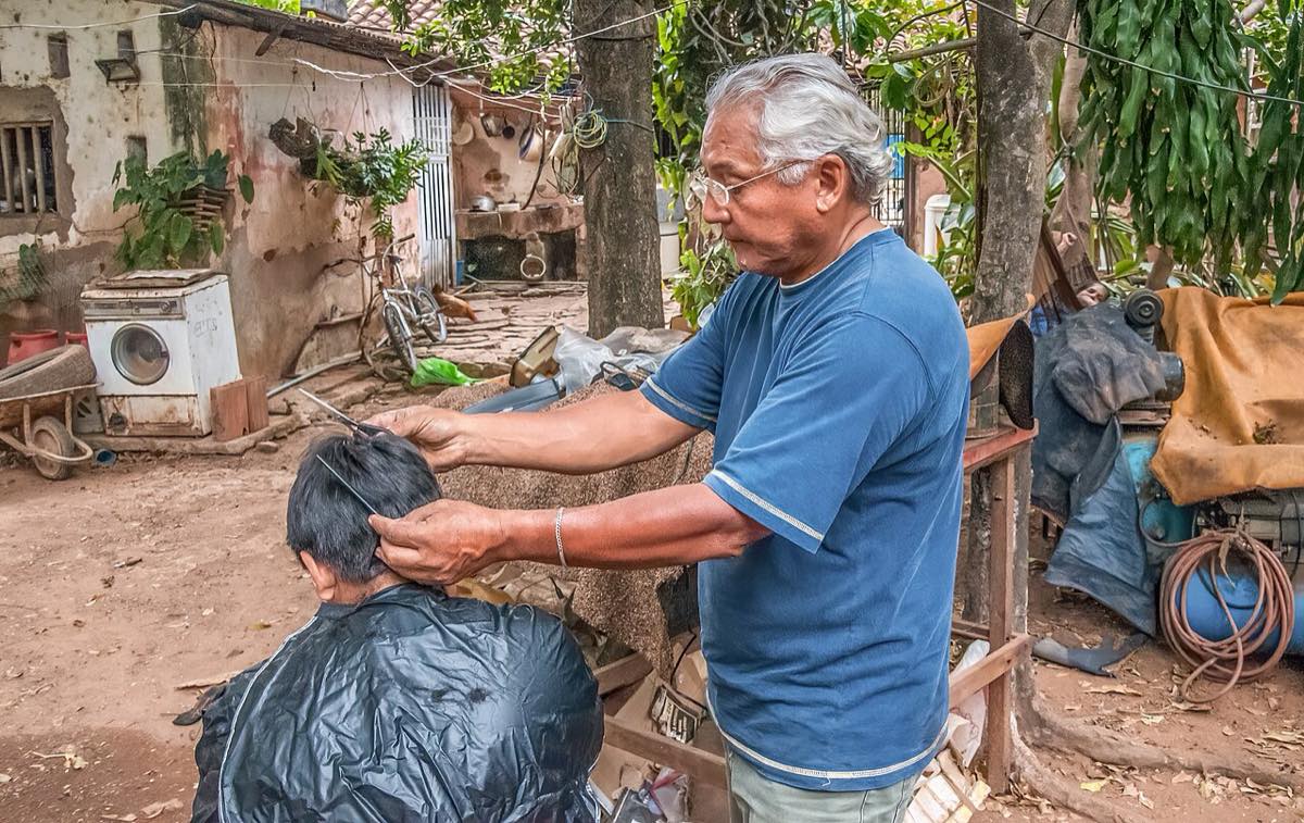 A gray-haired hairdresser in a blue T-shirt lifts a section of a seated customer's short dark hair with a comb and cuts it with scissors, outdoors