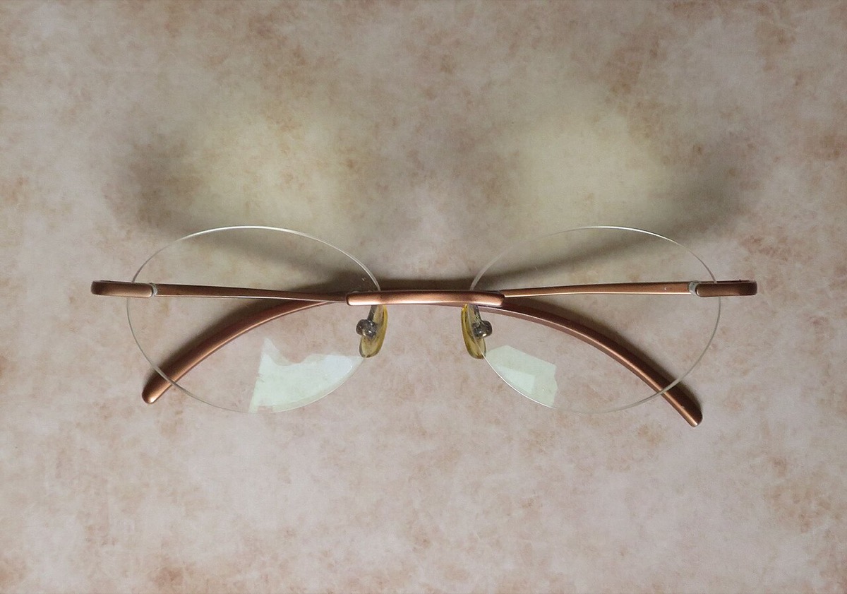 A pair of rimless eyeglasses with oval lenses and thin copper-colored metal arms, folded on a beige surface