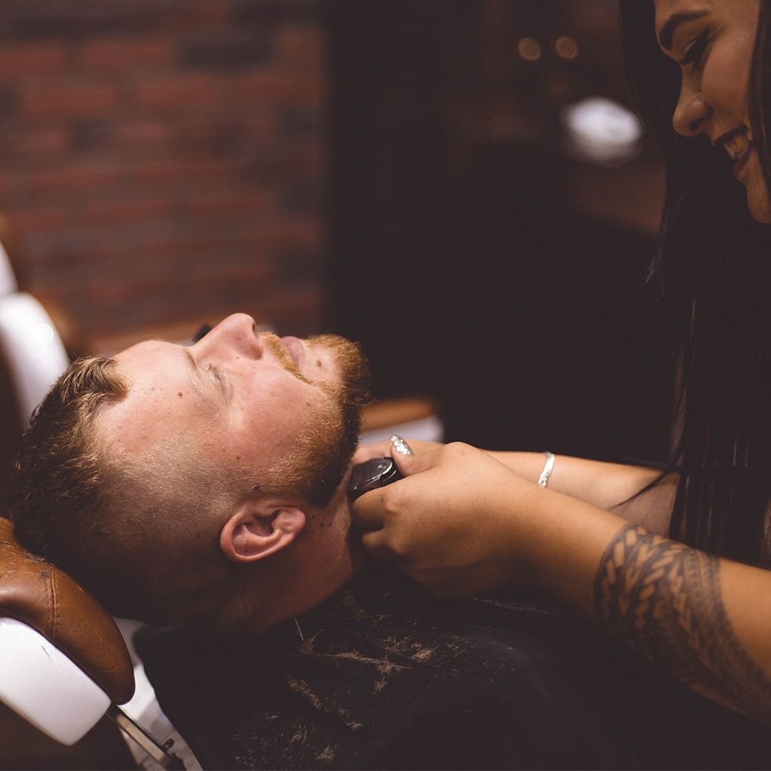 A barber uses electric clippers to trim a reclining client's short beard along the jaw and neck