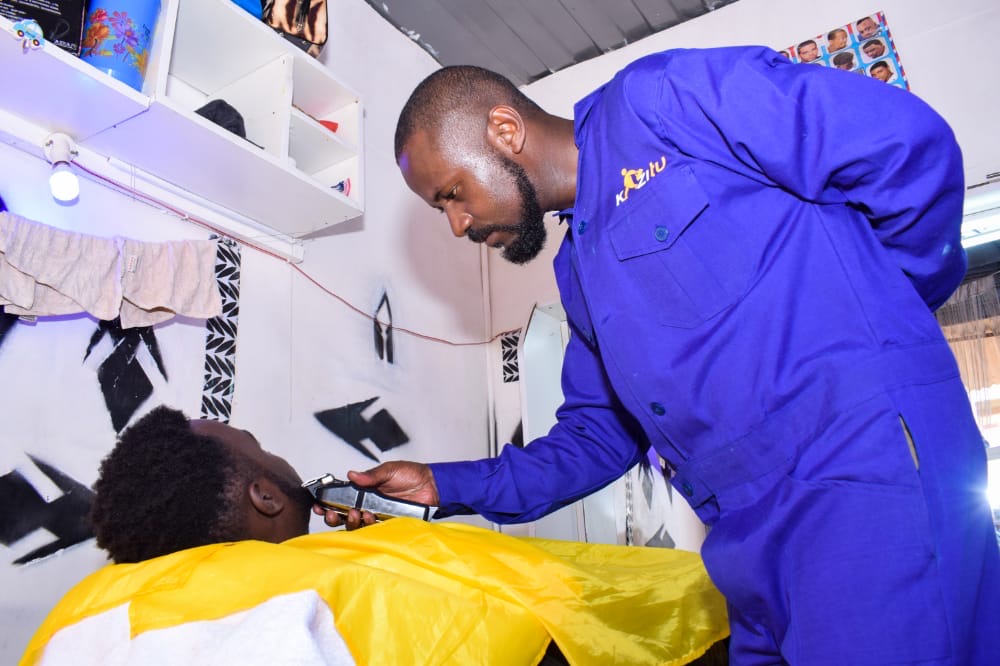A barber in a blue work suit trims a reclining customer's beard along the jaw with electric clippers in a barbershop in Kenya