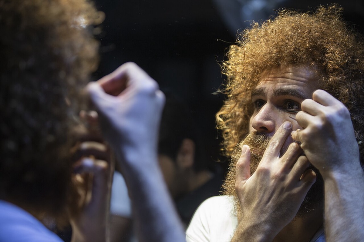 A man with curly red hair and a beard looks into a mirror while drawing kohl along his lower eyelid with a thin applicator