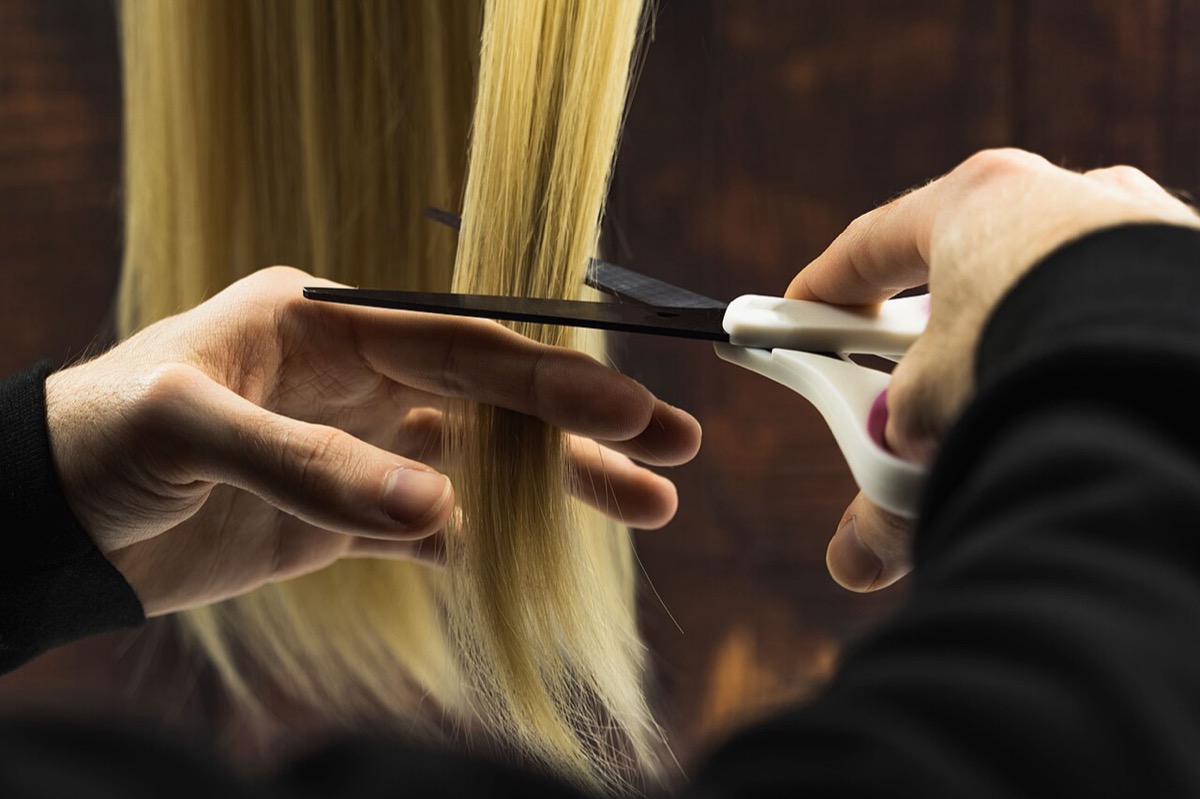 A hairdresser's hands holding a section of long straight blonde hair between two fingers and cutting it with scissors