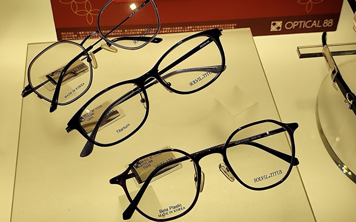 Three eyeglass frames on a shop display stand: a slim rectangular frame in the middle and two rounded geometric frames above and below it